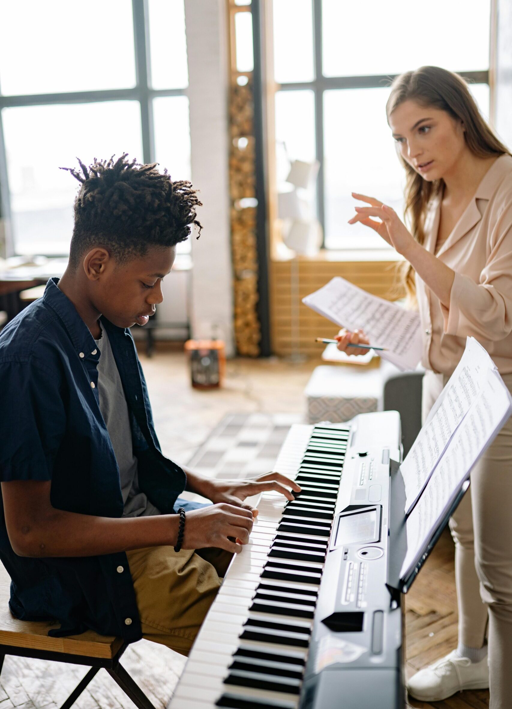 A teenage student plays an electric keyboard while a music teacher offers guidance and holds sheet music.