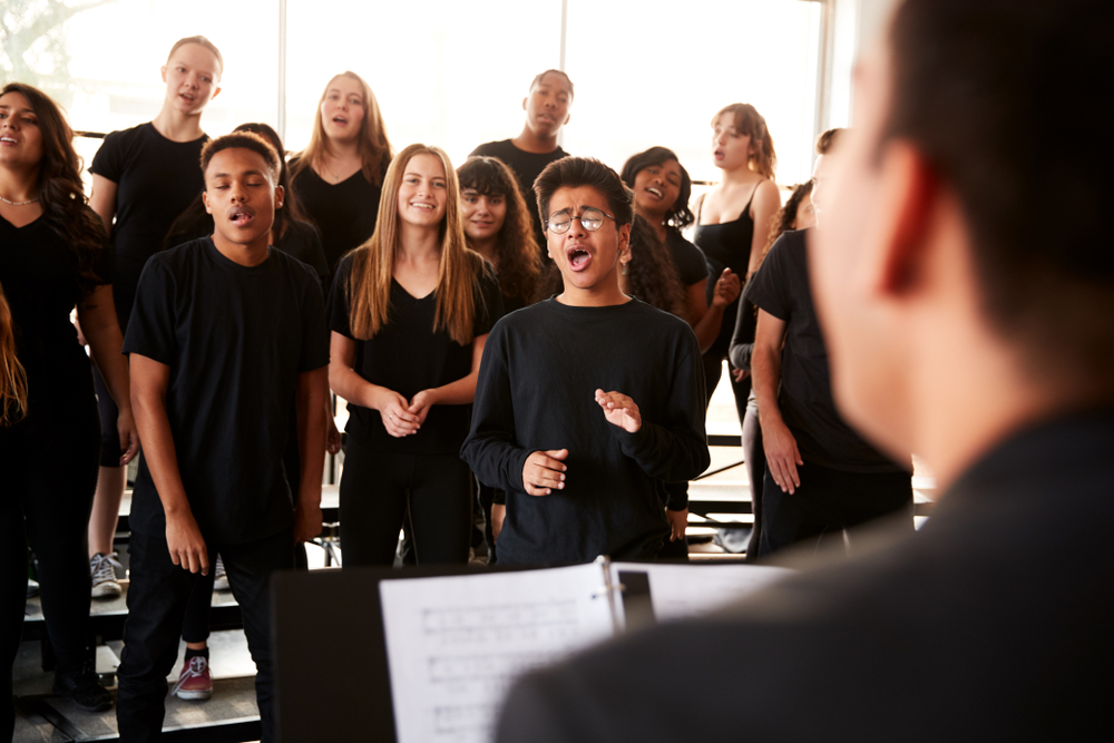 A diverse group of students performs in a choral rehearsal, guided by a conductor in a well-lit music classroom / performing arts school.