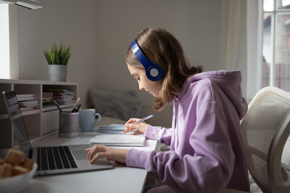 A focused student wears headphones while studying at a laptop, taking notes during an online lesson from home.
