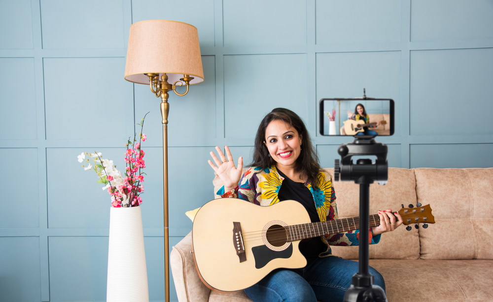 A cheerful guitar instructor records an online lesson or livestream from home, engaging directly with her virtual students.