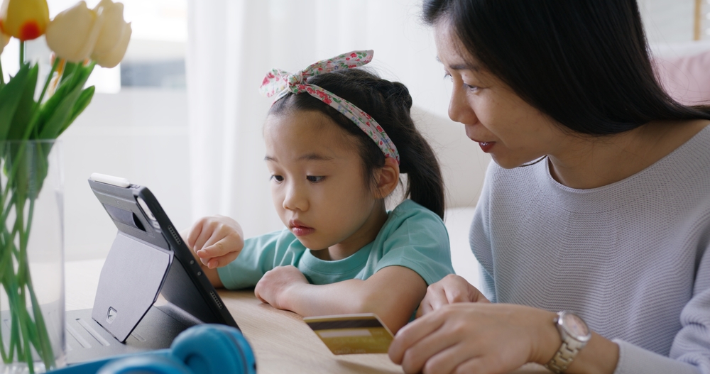 Young,Girl,Little,Kid,Help,Mom,Pay,School,Charge,Tuition A parent and child sit together at a tablet, navigating an online payment or enrollment form with a credit card in hand.