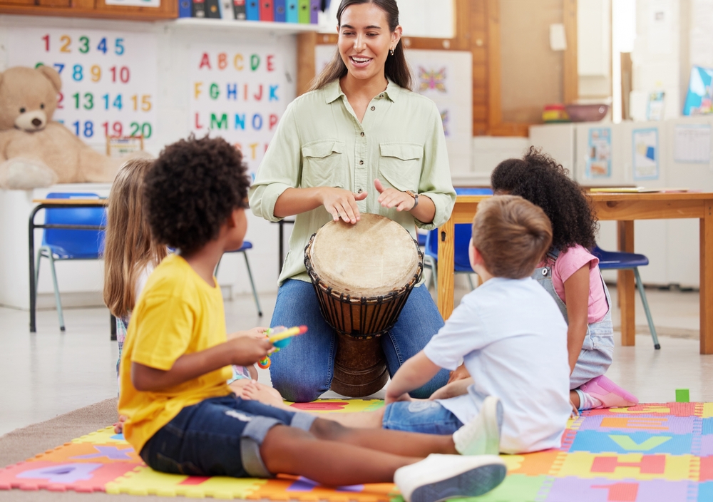 This,Is,How,You,Play,It.,Shot,Of,A,Woman A preschool music teacher leads a fun and interactive drumming circle with young children in a colorful classroom.