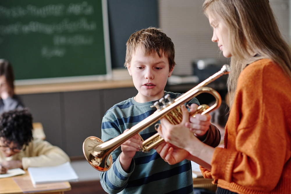 A music teacher helps a student learn finger placement during a trumpet lesson in a classroom setting.