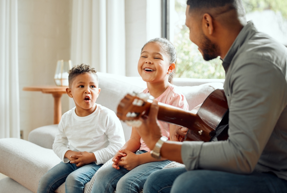 Two young children sing and laugh while their teacher plays guitar during a joyful music lesson at home.