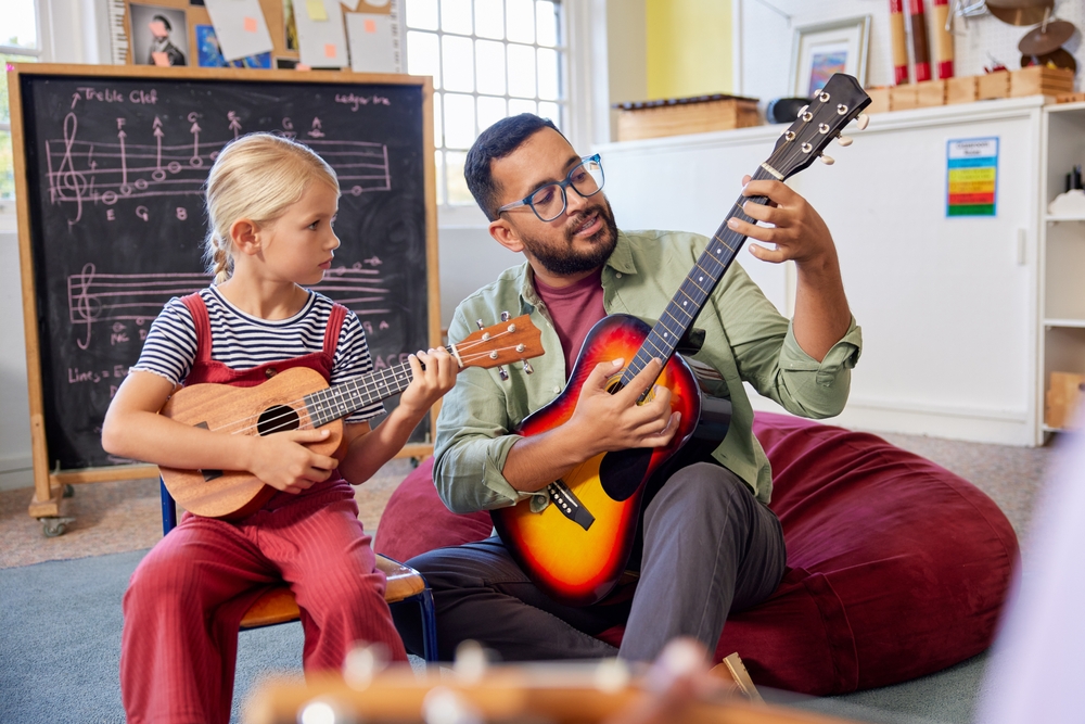 Man in glasses sitting on beanbag chair giving guitar lesson to young girl in a classroom