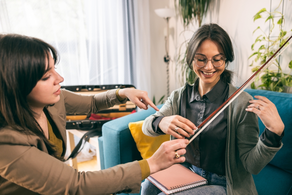 Music,Teacher,Having,Violin,Lesson,With,Student,While,She,Teaches An instructor and student share a hands-on violin lesson in a relaxed home environment, focusing on bow technique.