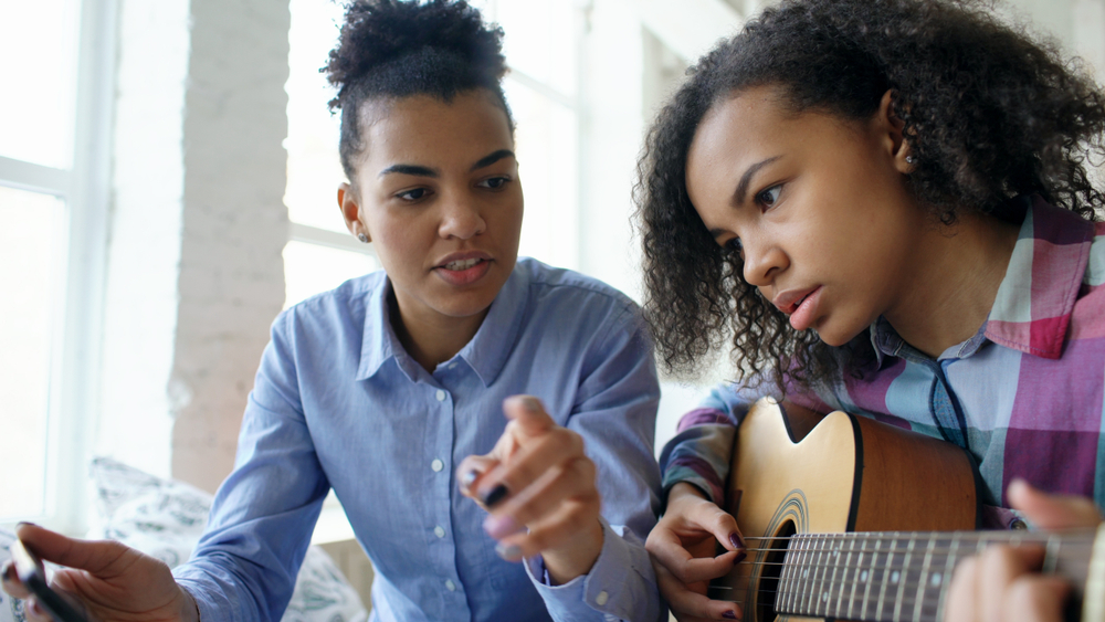 Young woman in blue shirt giving guitar lesson to a young girl, focusing intently